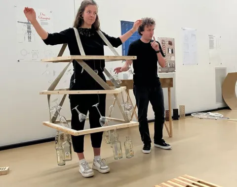 Zwei Studierende präsentieren ein kreatives Designprojekt im Rahmen des Advanced Studio Handwerk an der Universität Liechtenstein. Eine Person trägt eine Konstruktion aus Holzlatten und Glasflaschen. Im Hintergrund sind Plakate und Modelle zu sehen.