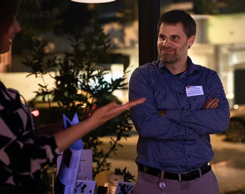 Eine Person steht lächelnd mit verschränkten Armen in einem Raum, während eine andere Person gestikuliert. Im Hintergrund sind Pflanzen und verschwommene Lichter zu sehen, was auf eine entspannte Atmosphäre bei einem Alumni-Treffen der Universität Liechtenstein hindeutet.