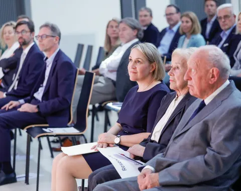 Eine Gruppe von Menschen sitzt in einem Auditorium. Im Vordergrund sitzen drei Personen in der ersten Reihe, aufmerksam nach vorne blickend. Die Veranstaltung scheint formell zu sein, mit mehreren Reihen von Teilnehmern im Hintergrund.