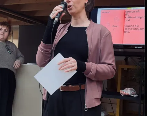 Eine Person hält ein Mikrofon und spricht vor einem Publikum. Sie trägt eine rosa Jacke und hält ein Blatt Papier. Im Hintergrund ist eine Präsentation auf einem Bildschirm zu sehen. Eine weitere Person steht links daneben und hört zu.