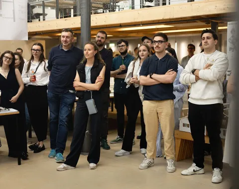 Eine Gruppe von Menschen steht in einem Innenraum der Universität Liechtenstein, aufmerksam einem Ereignis folgend. Im Hintergrund sind architektonische Modelle und eine Galerieebene zu sehen. Die Atmosphäre wirkt interessiert und gespannt.