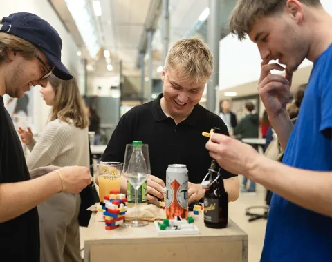Drei junge Erwachsene stehen um einen Tisch mit Getränken und Snacks, lachen und interagieren miteinander. Im Hintergrund sind weitere Personen in einer hellen, modernen Umgebung zu sehen.