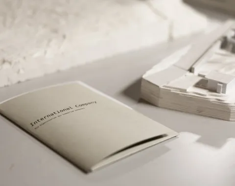 Ein Heft mit der Aufschrift "International Company" liegt auf einem Tisch neben einem Architekturmodell. Das Bild vermittelt eine professionelle und kreative Atmosphäre, passend für eine universitäre Umgebung.