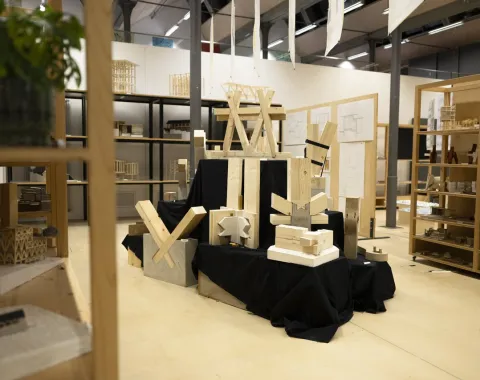 Ausstellung von Holzmodellen und Bauteilen in einem modernen Raum. Die Modelle sind auf schwarzen Stoffen arrangiert und zeigen verschiedene architektonische Strukturen. Im Hintergrund sind Regale mit weiteren Modellen und Zeichnungen sichtbar.
