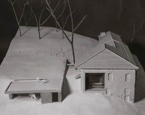 Ein Architekturmodell zeigt ein Gebäude mit Satteldach und angrenzendem offenen Unterstand auf einer schneebedeckten Landschaft. Im Hintergrund stehen stilisierte Bäume.