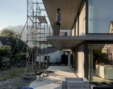 Ein Architekt steht auf einem modernen Gebäude mit großen Glasfenstern, daneben ein Baugerüst. Im Hintergrund sind Berge und ein Haus zu sehen. Die Szene zeigt eine Baustelle bei Sonnenuntergang.