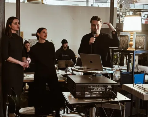 Eine Gruppe von Personen steht in einem Raum mit moderner Einrichtung. Eine Person hält ein Mikrofon und spricht, während andere zuhören. Im Hintergrund sind Tische, Stühle und technische Geräte zu sehen. Die Szene wirkt wie eine Präsentation oder ein Workshop in einem akademischen Umfeld.