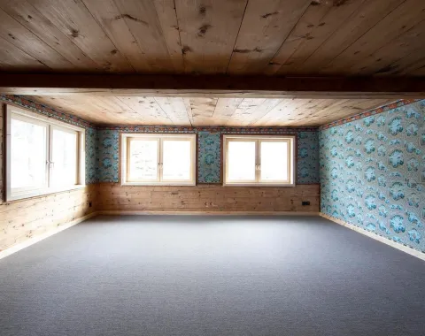 Ein leerer Raum mit Holzbalkendecke und Holzwänden, teilweise mit blauer, gemusterter Tapete. Drei große Fenster lassen Tageslicht herein. Der Boden ist mit grauem Teppich ausgelegt.