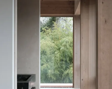 Innenraum mit Holzverkleidung und großem Fenster, das den Blick auf grüne Bäume freigibt. Moderne Architektur mit Fokus auf natürliche Materialien und Licht.