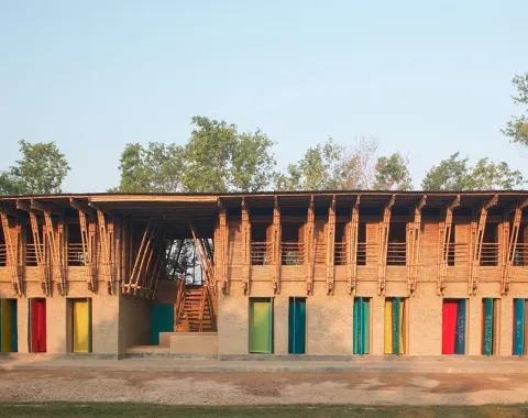 Ein zweistöckiges Gebäude aus natürlichen Materialien wie Lehm und Bambus. Die Fassade zeigt bunte Türen und Fenster, umgeben von Bäumen. Das Design kombiniert traditionelle Bauweisen mit modernem Stil.