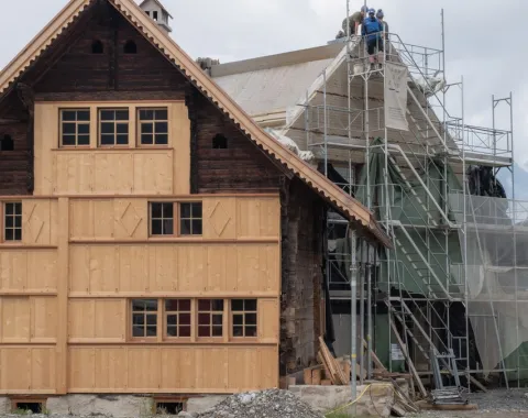 Ein traditionelles Gebäude wird renoviert. Links ist eine Holzfassade zu sehen, rechts stehen Gerüste mit Bauarbeitern auf dem Dach. Der Himmel ist bewölkt.