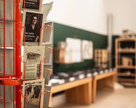 Ein roter Postkartenständer mit Architekturpostkarten im Vordergrund, im Hintergrund ein Raum mit Architekturmodellen und Zeichnungen auf einem Tisch.