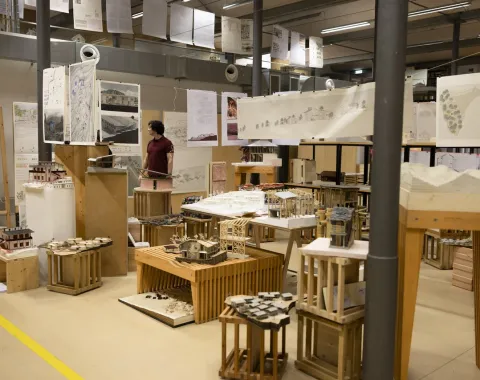Eine Ausstellung in einem Raum mit Architekturmodellen und Plänen. Eine Person betrachtet die ausgestellten Werke. Die Umgebung wirkt industriell und kreativ, mit zahlreichen Zeichnungen und Modellen auf Tischen und Podesten.