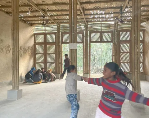 Kinder spielen und tanzen in einem neu gebauten Schulgebäude aus Bambus in Bihar, Indien. Im Hintergrund sitzen weitere Kinder auf dem Boden. Das Gebäude hat eine natürliche, lichtdurchflutete Atmosphäre.