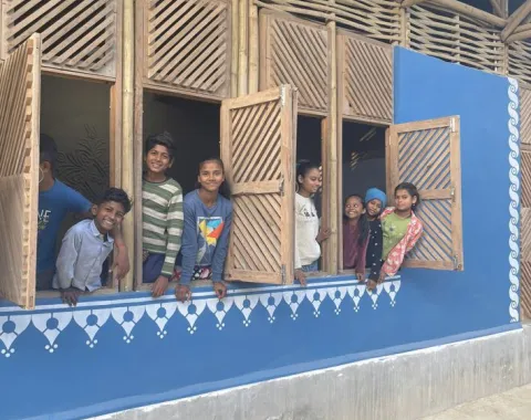 Kinder schauen lächelnd aus den Fenstern eines neu gebauten Schulgebäudes in Bihar, Indien. Die Fassade ist blau mit dekorativen weißen Mustern.