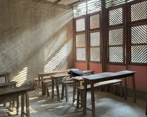 Ein einfacher Klassenraum mit Holztischen und -bänken, durchflutet von Sonnenlicht, das durch Fenster mit Holzlamellen fällt.