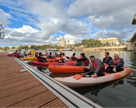 Studierende in Kanus auf einem Fluss in Sevilla, mit Schwimmwesten, vor einer malerischen Stadtlandschaft und bewölktem Himmel.