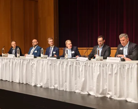 Eine Gruppe von sechs Personen sitzt an einem langen Tisch auf einer Bühne. Sie tragen formelle Kleidung und haben Namensschilder vor sich. Im Hintergrund sind Vorhänge zu sehen. Der Tisch ist mit einem weißen Tuch bedeckt und es stehen Wasserflaschen darauf.
