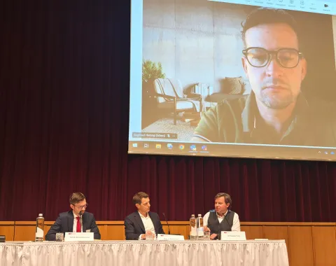 Ein Podium mit drei Männern, die an einem Tisch sitzen und diskutieren. Im Hintergrund ist ein großer Bildschirm mit dem Bild eines weiteren Mannes in einer Videokonferenz zu sehen. Die Szene findet in einem Konferenzsaal statt.