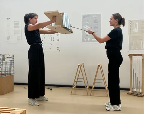 Zwei Studierende in einem Designstudio präsentieren ein Modell. Sie stehen sich gegenüber und halten gemeinsam eine Konstruktion aus Holz und Metall. Im Hintergrund sind Plakate mit architektonischen Entwürfen zu sehen.