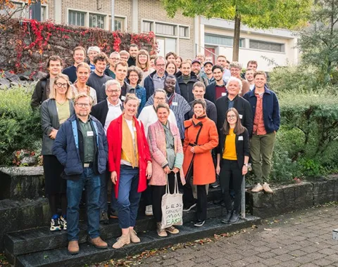 Gruppe von Menschen steht lächelnd auf einer Treppe im Freien, umgeben von grüner Vegetation. Sie tragen Namensschilder und sind für ein Symposium an der Universität Liechtenstein versammelt.