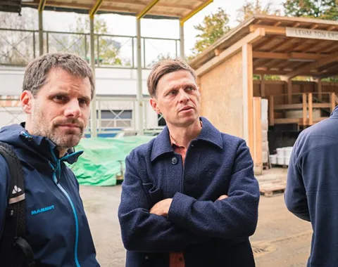 Zwei Männer stehen auf einer Baustelle, umgeben von Holzstrukturen und Baumaterialien. Sie tragen Jacken und schauen aufmerksam in die Ferne. Im Hintergrund sind Baugerüste und ein überdachter Bereich zu sehen.