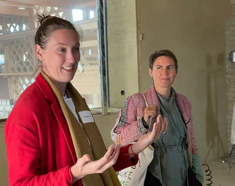 Zwei Personen stehen in einem Raum mit Lehmwänden. Eine Person trägt einen roten Blazer und gestikuliert, während die andere Person aufmerksam zuhört. Im Hintergrund sind Holzelemente sichtbar.