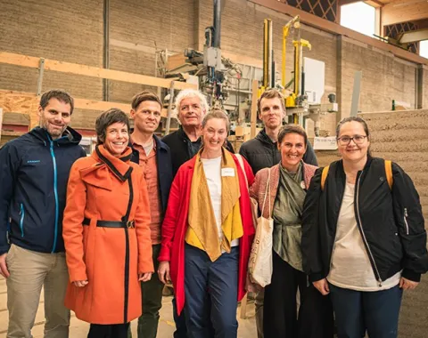 Eine Gruppe von acht Personen steht in einer Werkhalle vor einer Maschine und einem Stampflehm-Element. Sie lächeln in die Kamera.