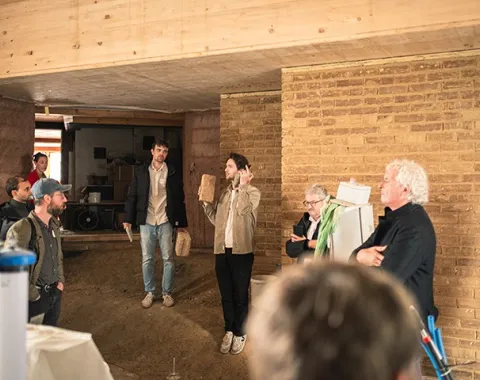 Eine Gruppe von Personen steht in einem Raum mit Wänden aus Stampflehm und Holz. Eine Person hält ein Präsentationsobjekt und spricht zu den anderen. Die Szene zeigt eine Diskussion über nachhaltige Baumaterialien bei einem Symposium der Liechtenstein School of Architecture.