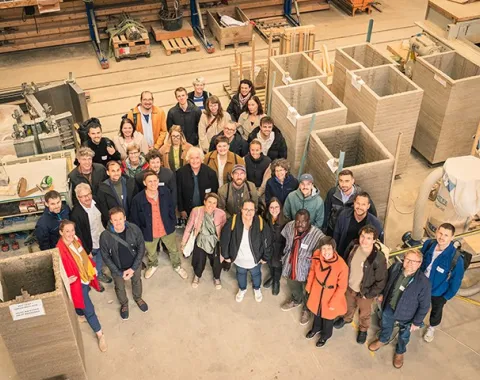 Gruppenfoto von Teilnehmern des Earth-Hub-Symposiums in einer Werkstatt, umgeben von Baumaterialien wie Stampflehm-Elementen.