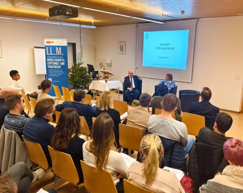 Ein Vortrag im Seminarraum der Universität Liechtenstein: Ein Redner und eine Rednerin sitzen vorne, während das Publikum aufmerksam zuhört. Im Hintergrund ist eine Präsentation mit dem Titel „Aus dem EFTA-Gerichtshof“ zu sehen. Ein Banner weist auf den LL.M.-Studiengang hin.