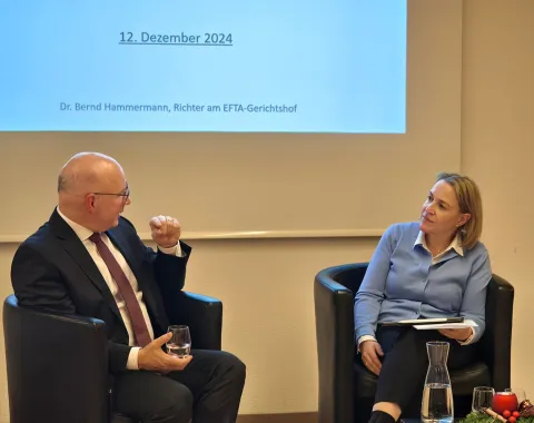 Zwei Personen sitzen in Sesseln vor einer Leinwand, die das Datum "12. Dezember 2024" zeigt. Eine Person hält ein Glas Wasser, die andere hat ein Notizbuch auf dem Schoß. Im Vordergrund steht eine Karaffe auf einem Tisch mit weihnachtlicher Dekoration.