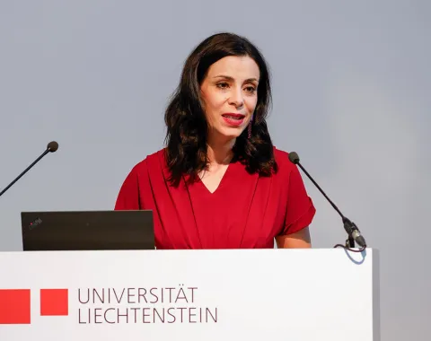 Eine Frau in rotem Kleid spricht an einem Rednerpult mit dem Logo der Universität Liechtenstein. Sie hält eine Rede vor einem Mikrofon.