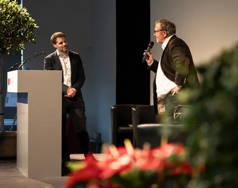 Zwei Männer stehen auf einer Bühne bei einer Veranstaltung. Einer spricht in ein Mikrofon, während der andere an einem Rednerpult steht. Im Hintergrund sind Pflanzen und ein großer Bildschirm zu sehen.