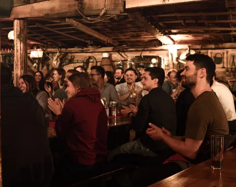 Eine Gruppe von Menschen sitzt in einem rustikalen Raum und applaudiert während einer Veranstaltung. Die Atmosphäre ist gemütlich und gesellig.