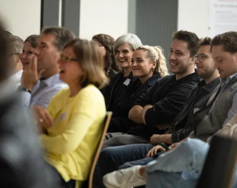 Eine Gruppe von Menschen sitzt in einem Auditorium und lächelt während einer Veranstaltung an der Universität Liechtenstein. Die Atmosphäre wirkt entspannt und fröhlich.