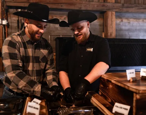Zwei Personen in Cowboyhüten bereiten Essen an einem Buffet vor. Sie tragen schwarze Handschuhe und stehen in einem rustikalen Raum mit Holzwänden. Vor ihnen sind Schilder mit verschiedenen Fleischsorten zu sehen.