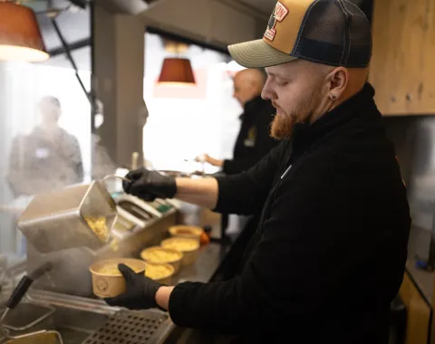 Ein Koch füllt dampfende Nudeln in Schalen in einem Foodtruck. Im Hintergrund sind weitere Personen und Küchenutensilien zu sehen.