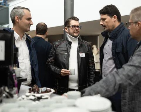 Eine Gruppe von vier Männern steht an einem Stehtisch und unterhält sich bei einer Veranstaltung. Einer hält eine Tasse Kaffee. Im Vordergrund sind Geschirr und Snacks zu sehen. Die Atmosphäre wirkt gesellig und professionell.