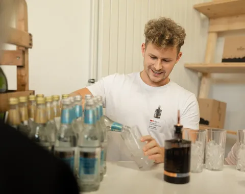 Ein junger Mann schenkt an einem Tisch Getränke ein. Auf dem Tisch stehen mehrere Flaschen und Gläser. Im Hintergrund sind Regale mit weiteren Gegenständen zu sehen.