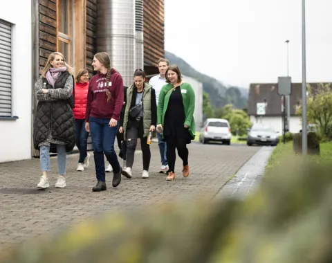 Eine Gruppe von Menschen geht auf einem gepflasterten Weg auf dem Campus der Universität Liechtenstein. Im Hintergrund sind Gebäude und eine bergige Landschaft zu sehen.