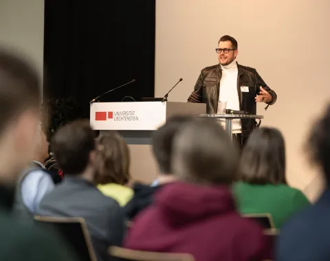 Ein Redner mit schwarzer Lederjacke steht auf der Bühne vor dem Redepult und hält eine Rede. Im vorderen Teil des Bildes ist ein Teil des Publikums zu sehen.