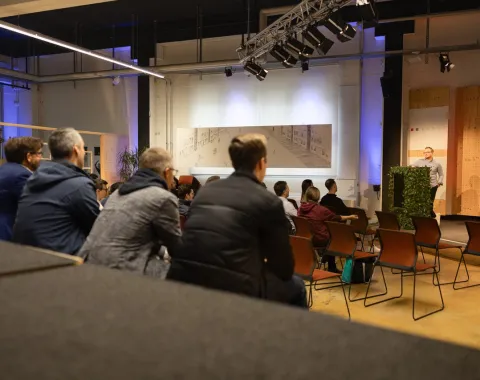Eine Gruppe von Menschen sitzt in einem Veranstaltungsraum und hört einem Vortrag zu. Im Hintergrund ist ein Redner am Podium zu sehen. Der Raum ist modern gestaltet und gut beleuchtet.