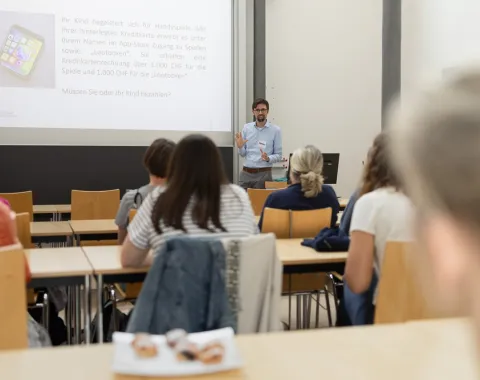Ein Dozent hält einen Vortrag in einem Seminarraum vor mehreren Erwachsenen. Die Zuhörer sitzen an Tischen und schauen zur Präsentation an der Leinwand.