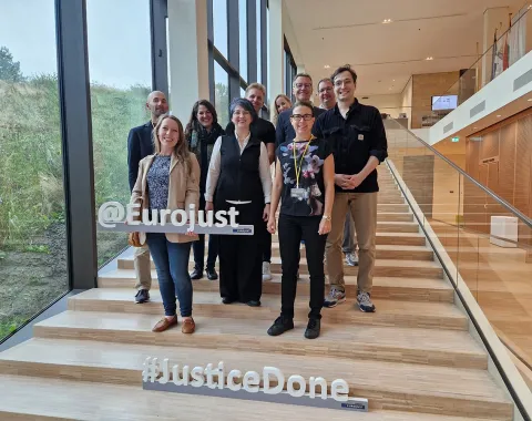 Gruppe von Studierenden und Dozenten des Executive Master of Laws (LL.M.) im Wirtschaftsstrafrecht posiert auf einer Treppe bei Eurojust in Den Haag. Im Vordergrund sind die Schriftzüge "@Eurojust" und "#JusticeDone" zu sehen.