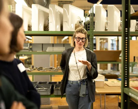 Eine Person steht in einem Lagerraum vor Regalen mit Bauteilen und spricht zu einer anderen Person. Die Szene findet während einer Exkursion zum Thema zirkuläres Bauen statt.