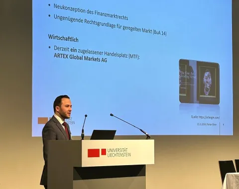 Ein Vortragender steht an einem Rednerpult der Universität Liechtenstein und präsentiert eine Folie mit dem Titel "Hintergrund". Die Folie enthält Informationen zur rechtlichen und wirtschaftlichen Neukonzeption des Finanzmarktrechts.