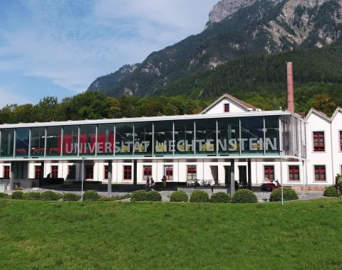 Außenansicht der Universität Liechtenstein mit modernem Glasgebäude und traditionellem weißen Gebäude im Hintergrund, umgeben von grüner Landschaft und Bergen.