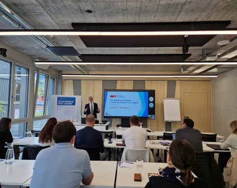 Ein Dozent hält einen Vortrag über Geldwäscherei im 20. und 21. Jahrhundert vor einer Gruppe von Teilnehmenden in einem modernen Seminarraum der Universität Liechtenstein. Im Hintergrund ist eine Präsentation auf einem Bildschirm zu sehen.