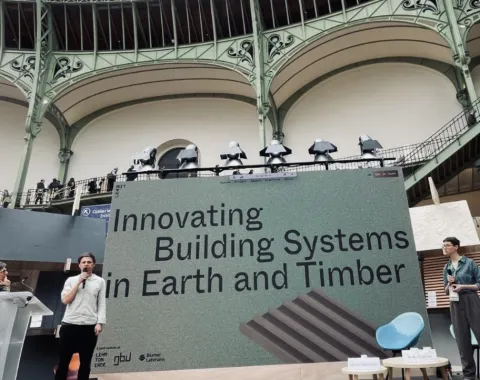 Ein Redner präsentiert auf einer Bühne vor einem großen Bildschirm mit der Aufschrift „Innovating Building Systems in Earth and Timber“ im Grand Palais, Paris. Im Hintergrund ist die beeindruckende Architektur des Veranstaltungsortes zu sehen.